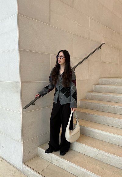 Mujer con cabello largo y gafas, vestida con suéter gris de rombos y pantalones negros, de pie en una escalera de mármol sosteniendo un bolso blanco.