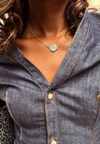 Kultasävyinen kaulakoru, jossa suorakaiteenmuotoinen aqua sininen riipus, puettuna tummansinisen farkkupaidan päälle, jossa on kultaiset tikkaukset ja napit.