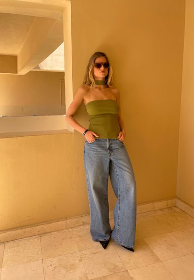 Top sin tirantes de rayas verdes, jeans de mezclilla anchos en azul claro y tacones negros de punta. La modelo lleva gafas de sol marrones y está de pie contra una pared beige.