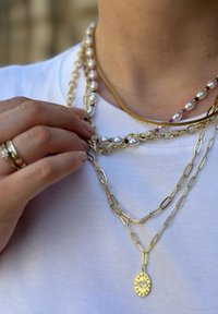 Drei geschichtete Halsketten: eine goldene Kette mit Gliedern, eine Perlenkette mit bunten Perlen und ein goldener Anhänger mit einem Edelsteinakzent.