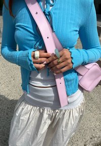 Ανοιχτό μπλε ριγέ κάρντιγκαν με κλείσιμο τύπου δέσιμο, τοποθετημένο πάνω από ένα λευκό τοπ. Ροζ δερμάτινη τσάντα με υφή. Λευκή φούστα που ρέει, δαχτυλίδια και τέχνη νυχιών.