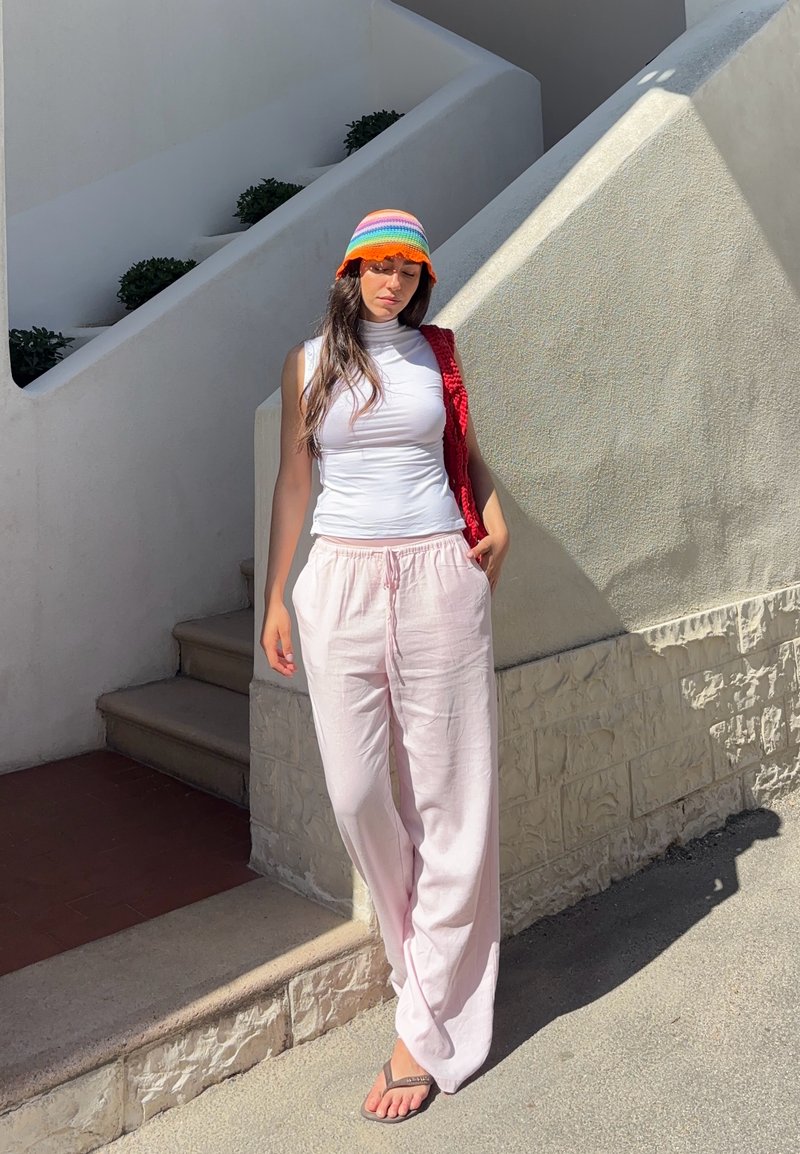 Femme portant un débardeur blanc, un pantalon rose clair, un chapeau arc-en-ciel et des sandales, debout contre un mur blanc texturé près d'escaliers extérieurs en plein soleil.