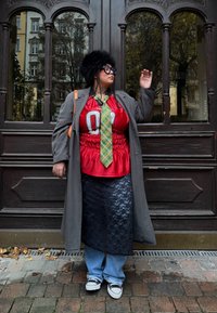 Červené tričko s kruhovou grafikou, zelená károvaná kravata, sivý trenčkot, čierna čipkovaná sukňa, voľné modré rifle a čierno-biele tenisky. Hnedé drevené pozadie.