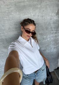 Top felpa bianca con zip e maniche corte, shorts in denim chiaro, occhiali da sole neri e borsa nera. Il tessuto morbido e la vestibilità rilassata creano un look informale.