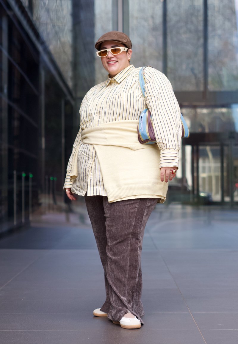 Personne portant une chemise rayée, un pull beige noué autour de la taille, un pantalon marron, des chaussures blanches, une casquette marron, des lunettes de soleil blanches, et tenant un sac à main bleu.