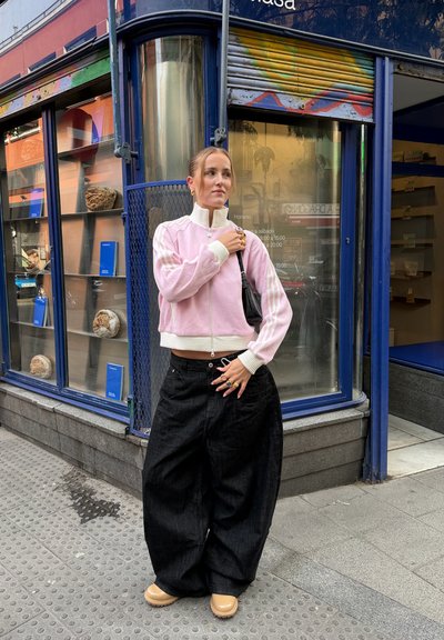 Chaqueta rosa recortada con cremallera y detalles blancos, pantalones de mezclilla negros extragrandes y zapatos beige sin cordones. Joyería que incluye anillos y un bolso de mano.