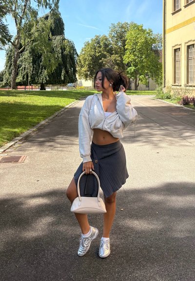 Modelo con un hoodie corto gris, falda plisada azul marino, zapatillas de deporte plateadas y sosteniendo un bolso crema. Al aire libre, con vegetación y edificios al fondo.