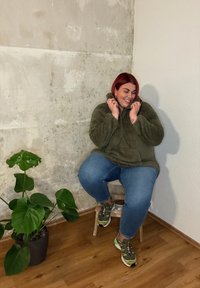 Chaqueta de piel de color verde oliva, vaqueros azules y zapatillas verdes. El modelo se sienta en un taburete de madera cerca de una planta en maceta contra una pared texturizada.