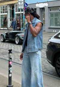 Chaleco de denim, camisa negra, pantalones de denim de pierna ancha, los accesorios incluyen un pañuelo y gafas de sol, de pie en una calle de adoquines.