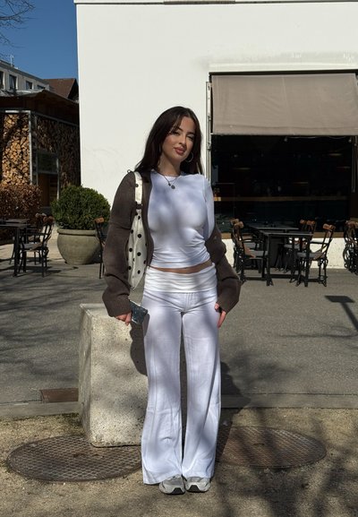 Joven de pie al aire libre cerca de asientos de cafetería, vestida con un conjunto blanco, un cárdigan marrón y sosteniendo un teléfono inteligente con un bolso de lunares en el hombro.