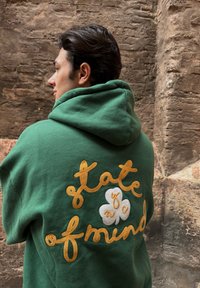 Green hooded sweatshirt featuring yellow and white embroidered text "state of mind" along with a floral design on the back. Made from soft fabric with a relaxed fit.