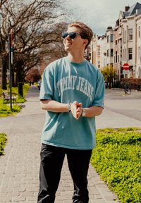 Šviesiai mėlyna medvilninė marškinėliai su "TOMMY JEANS" užrašu, juodos kelnės, tamsūs saulės akiniai, stovintis ant akmenimis grįsto tako su žaluma.