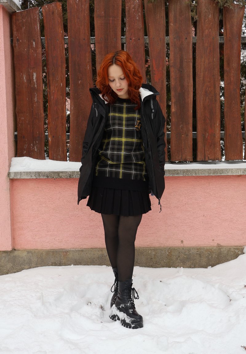 Jeune femme aux cheveux roux portant une veste noire, un pull à carreaux, une jupe noire et des bottes, debout sur la neige devant une clôture en bois et un mur rose.