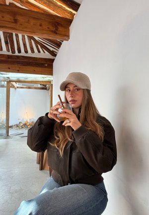 Joven mujer con chaqueta marrón y gorra beige sentada en interiores, sosteniendo una bebida con pajita cerca de su boca, con vigas de madera en el techo.