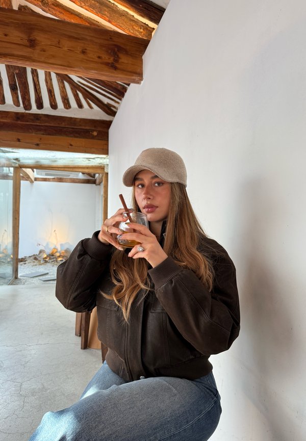 Junge Frau in einer braunen Jacke und beiger Kappe sitzt drinnen und hält ein Getränk mit einem Strohhalm nahe am Mund, darunter Holzbalken.