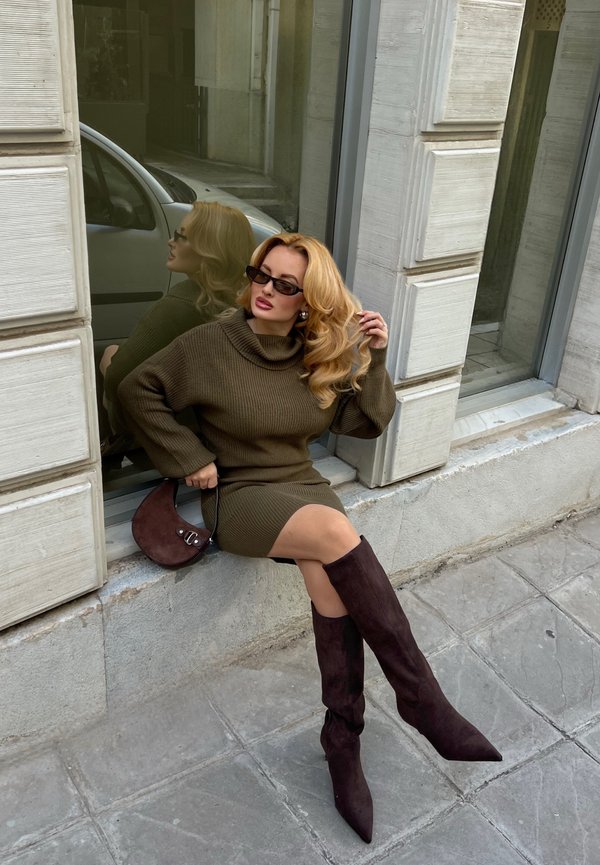 Kvinde med lyst hår iført mørke solbriller, en brun sweaterkjole og knælange støvler, der sidder på en stenkant ved et vindue.