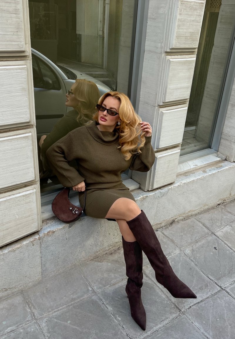 Femme aux cheveux blonds portant des lunettes de soleil foncées, une robe-pull marron et des bottes montant jusqu'aux genoux, assise sur un rebord en pierre près d'une fenêtre.