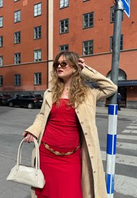 Beige velúr trench kabát vörös felső és hozzá illő szoknya felett, aranylánc öv, fekete napszem�üveg és fehér táska. Vörös téglaépületek a háttérben.