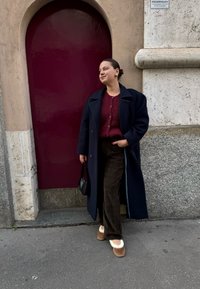 Marinblå kappa över en vinröd stickad tröja; bruna rutig byxor; beige och vita tofflor; svart väska; står vid en vinröd bågformad dörr.