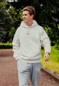 Ανοιχτόχρωμη φούτερ με κουκούλα με μικρό κόκκινο λογότυπο Nike, συνδυασμένη με γκρι φόρμες που διαθέτουν μια διακριτική λογότυπο Nike. Εξωτερικό περιβάλλον με πράσινη βλάστηση.