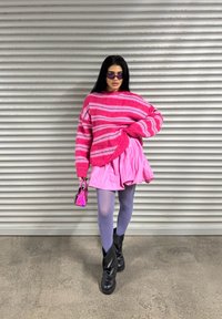 Suéter oversized riscado em rosa, usado sobre uma saia leve rosa. O conjunto inclui meias roxas e botas pretas de tornozelo, com uma pequena bolsa rosa.