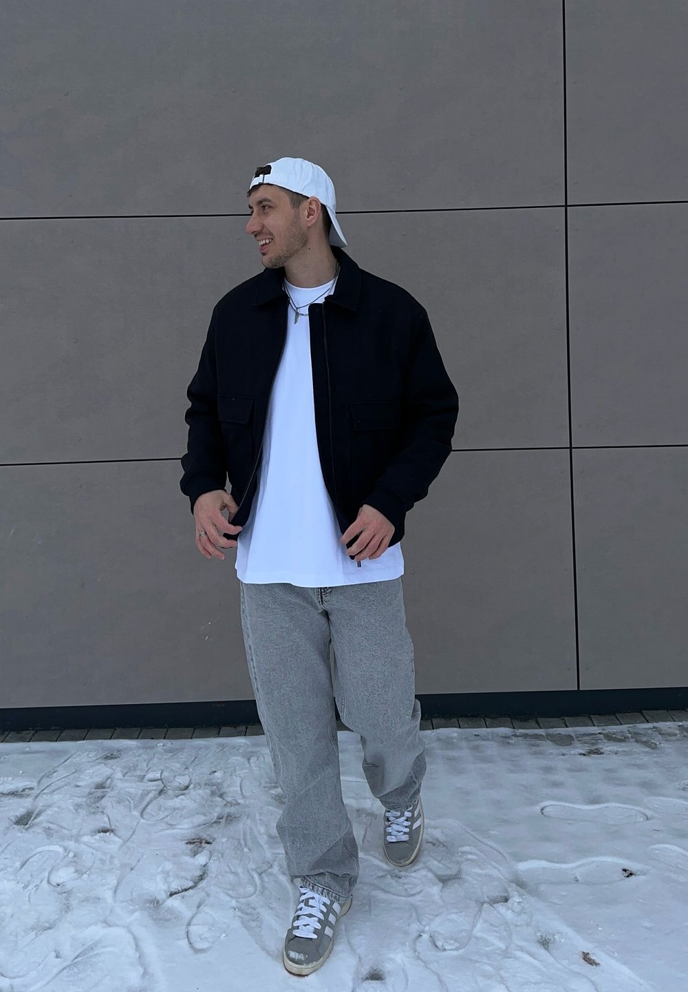 Jeune homme marchant sur la neige, portant une casquette blanche à l'envers, une veste noire, un t-shirt blanc, un pantalon gris et des baskets, devant un mur gris.