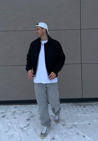 Jeune homme marchant sur la neige, portant une casquette blanche à l'envers, une veste noire, un t-shirt blanc, un pantalon gris et des baskets, devant un mur gris.