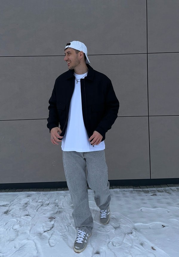 Jeune homme marchant sur la neige, portant une casquette blanche à l'envers, une veste noire, un t-shirt blanc, un pantalon gris et des baskets, devant un mur gris.