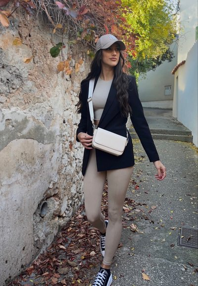 Blazer negro, top corto beige, leggings de canalé beige y zapatillas negras. Bolso cruzado beige. El fondo presenta una pared de piedra y hojas caídas.