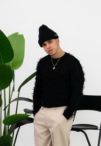 Pulover negru pufos cu margini destrămate, pantaloni bej și o căciulă neagră. Frunze verzi în fundal, inclusiv un scaun minimalist.