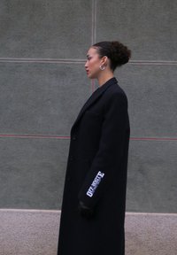 Profil d'une femme avec un chignon bouclé portant de grandes boucles d'oreilles en créole et un long manteau noir avec un texte blanc sur la manche, debout contre un mur gris.