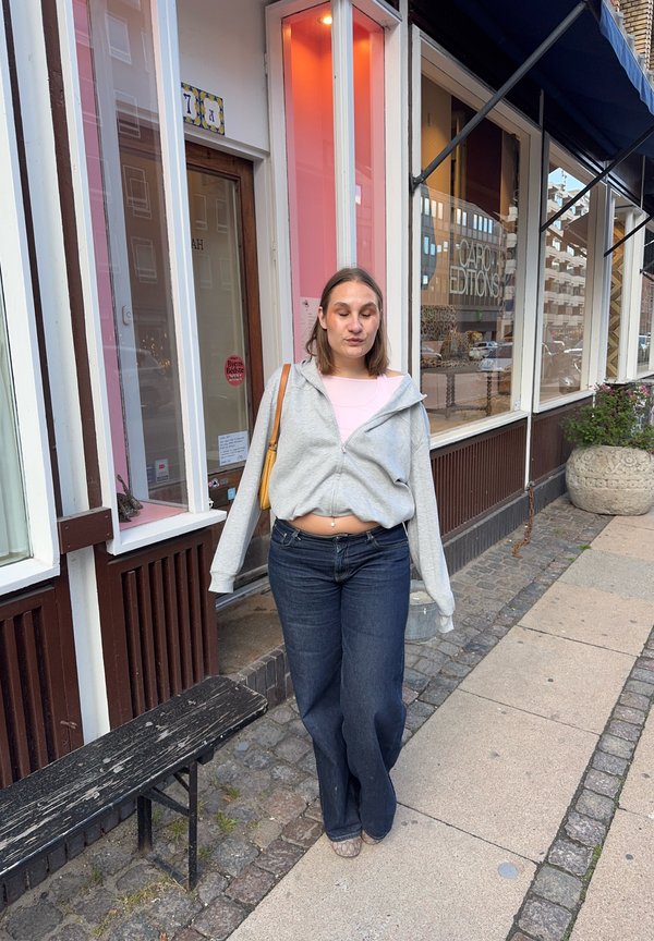 Mujer con sudadera gris y vaqueros azules caminando por una acera de la ciudad junto a escaparates y un banco de madera.