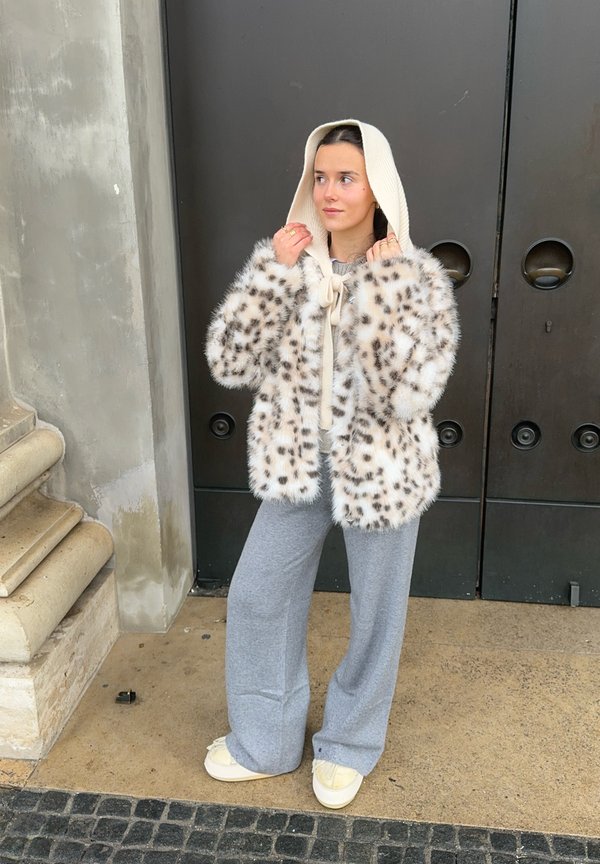 Palton din blană artificială cu pete de leopard maro, glugă crem, legat la gât. Purta pantaloni largi gri și pantofi bej, stând pe o suprafață de piatră.
