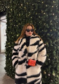 Une femme portant un manteau en fausse fourrure rayé noir et blanc et des lunettes de soleil se tient devant un grand sapin de Noël décoré de lumières blanches.