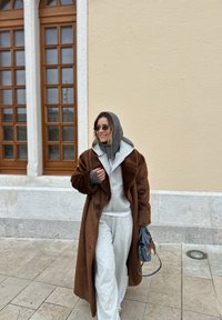 Femme marchant à l'extérieur portant un manteau en fourrure marron par-dessus un sweat à capuche gris et un pantalon de survêtement, portant un sac à main bleu, près d'un bâtiment beige avec de grandes fenêtres.