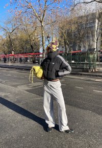 Personne portant une veste Adidas noire, un pantalon gris, un bonnet à motifs, tenant un sac jaune, debout dans une rue de la ville avec des arbres sans feuilles sous le soleil.