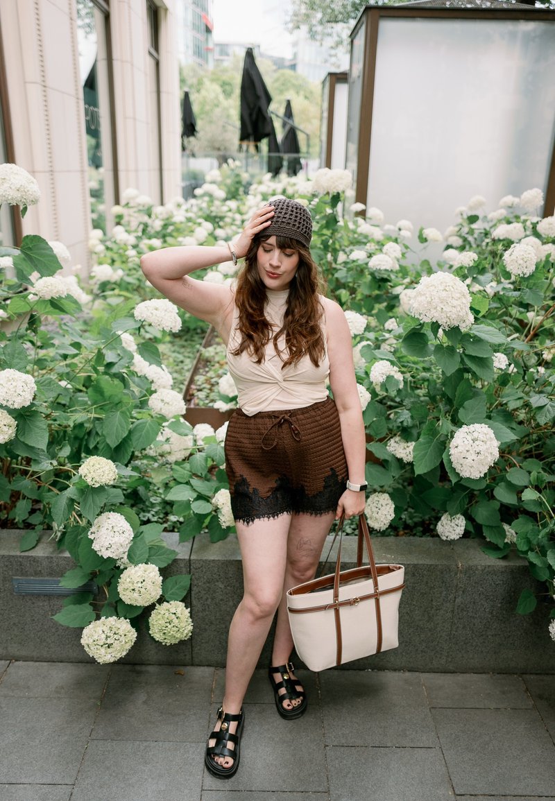 Jeune femme en shorts marron et bonnet tricoté se tient près d'hortensias blancs, tenant un sac à main beige à l'extérieur sur un chemin pavé.