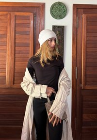Cappotto trench light cream su una maglietta nera, pantaloni neri e un berretto bianco a coste; tiene in mano una piccola borsa beige a fantasia.