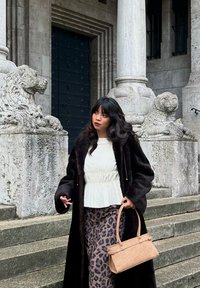 Mørk pelsjakke over en hvit peplumtopp og en brun leopardmønstret skjørt. Holder en lys brun, strukturert håndveske. Statuene av steinløver i bakgrunnen.