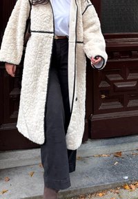 Casaco texturizado de cor creme com piping preto, featuring mangas largas e fecho com botões, combinado com calças pretas e uma camisa branca.