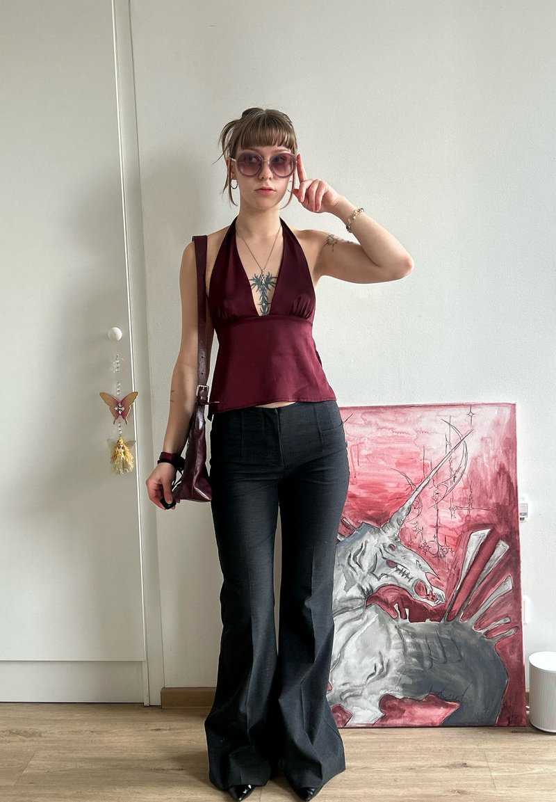 Tânără purtând ochelari de soare, un top halter burgundy și pantaloni evazați negri, stă în interior lângă un perete cu o pictură roșie și gri a unui unicorn.