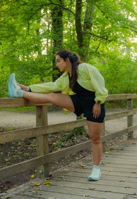 O femeie îmbrăcată într-o jachetă galbenă și pantaloni scurți negri își întinde o mostră de picior pe o balustradă din lemn într-o pădure verde, stând pe un drum din lemn acoperit cu frunze.