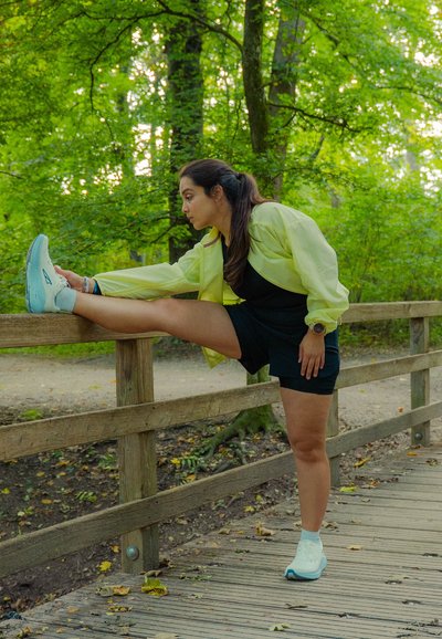 Mujer con chaqueta amarilla y pantalones cortos negros estira una pierna sobre una barandilla de madera en un bosque verde, de pie sobre un camino de madera cubierto de hojas.