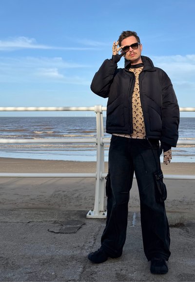 Chaqueta pespunteada negra con cuello alto, camisa beige de manga larga con patrón, pantalones negros estilo cargo y zapatos negros. Diseño de rebanadas de luna visible.