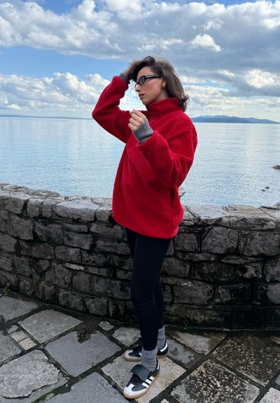 Sudadera de forro polar rojo con cuello alto, puños grises, leggings negros y zapatillas negras y doradas con calcetas grises, de pie cerca de un muro de piedra junto al agua.