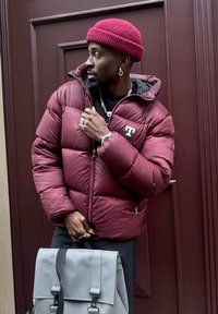 Casaco puffer bordeaux com fecho éclair e capuz, com acessórios em prata, usado sobre uma camisa preta. Mochila cinza com aba e alças.