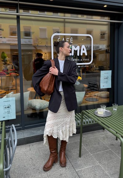 Una mujer vestida con una chaqueta oscura de rayas sobre una camisa blanca, una falda de encaje crema y botas marrones hasta la rodilla está de pie frente a un café.