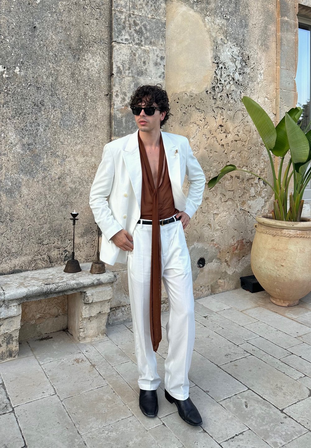 Uomo vestito con un completo bianco, camicia marrone con una lunga sciarpa, stivali neri e occhiali da sole, in piedi su un patio di pietra accanto a un muro rustico e una grande pianta in vaso.