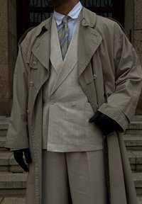 Μπεζ παλτό σε στυλ trench coat πάνω από ένα ανοιχτό λινό γιλέκο διπλής όψης, συνδυασμένο με ανοιχτούς παντελόνες; τονίζεται από μία ριγέ γραβάτα και μαύρα γάντια.