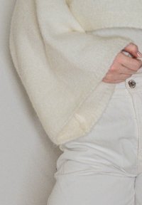 Sweater creme de corte solto com mangas bufantes, feito de um tecido de textura suave, usado sobre calças brancas de cintura alta com um botão metálico.